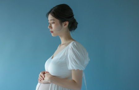Young pregnant woman in a white dress touching her belly isolated on a blue background.の素材