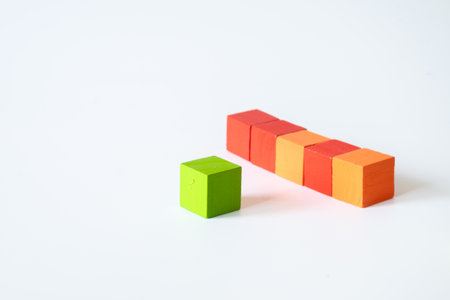 A single green wooden cube placed apart from a line of four stacked orange and red cubes on a white background, representing separation and creative contrast.の写真素材