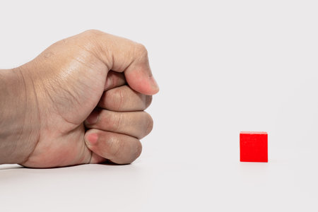 Human fist positioned near a red wooden block on a white background, illustrating concept of strength, determination, or challenge.の写真素材