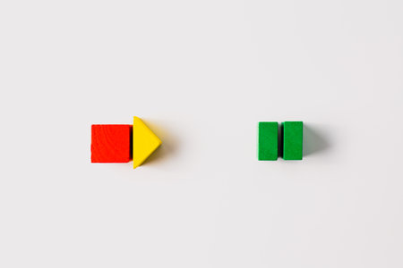 Red wooden cube and yellow triangle arranged as a rightward arrow, positioned opposite two green rectangular blocks on a clean white background in a creative minimal layout.の写真素材