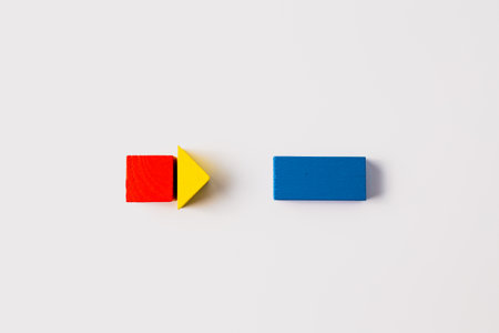 Minimalist composition featuring a red cube and yellow triangle forming a rightward arrow, pointing towards a single blue rectangular block on a clean white background.の写真素材