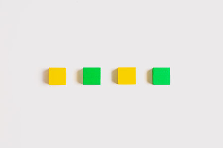 Four small wooden cubes, alternating between yellow and green, arranged in a neat horizontal line on a clean white background.の写真素材
