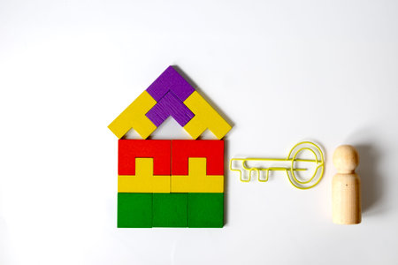 Colorful house-shaped geometric blocks, a yellow key-shaped paper clip, and a single wooden peg doll on a white background symbolizing home ownership, individuality, and opportunity.の写真素材