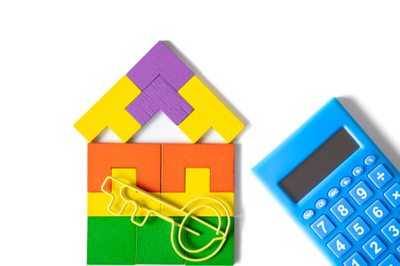 Colorful house-shaped wooden blocks with a yellow key-shaped paperclip and blue calculator on a white background, symbolizing home ownership, investment, and financial planning.の写真素材