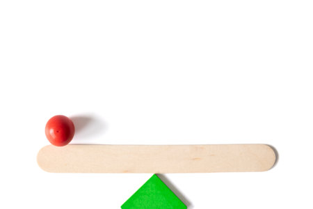 Minimalist image of a wooden stick balanced on a green triangle with a single red sphere on one side, symbolizing imbalance, decision-making, and risk.の写真素材