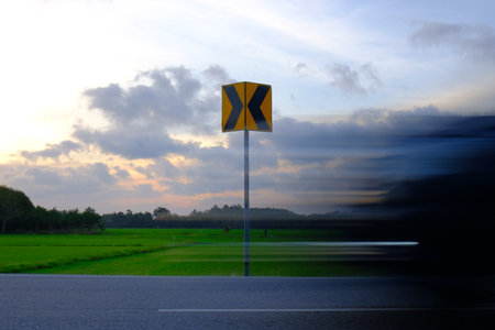 A blurred vehicle speeds past a yellow chevron road sign among green paddy fields at dusk, symbolizing direction and the momentum of fast decision-making.の写真素材
