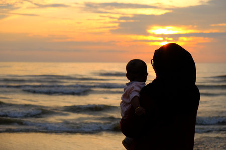 A mother wearing a black hijab holds her baby at the beach, watching the sunset over gentle waves, portraying warmth and family bonding.の写真素材