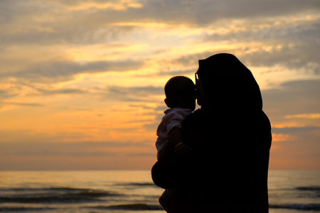 A mother in hijab tenderly kisses her baby by the beach at sunset, emphasizing love and family warmth in a beautiful natural setting.の写真素材