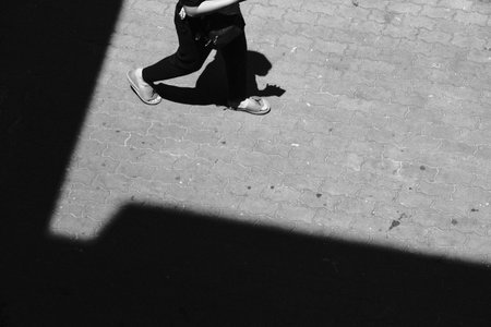 Monochrome, high-angle street photo showing a person's feet walking on patterned pavement, cut by a sharp, geometric shadow.の写真素材