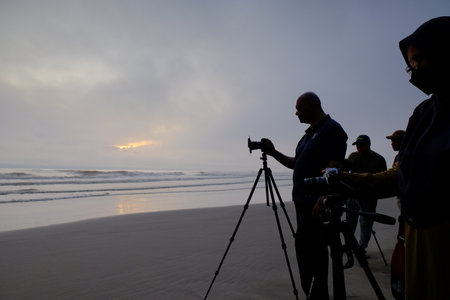 Photographers setting up cameras on tripods on a vast sandy beach, capturing the sun's first light breaking through the clouds over the sea.の写真素材
