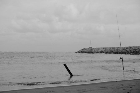 Black and white image of calm sea, rocky breakwater, lighthouse, and fishing rod on the beach under...