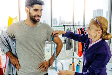 Creative Garment Design Tailoring Service Concept. Cute Professional Sewer Measuring Male Bearded Model Waist or Hip. Smiling Outfit Designer Taking Man Measurement for Formal Suit with Tape Meterの写真素材