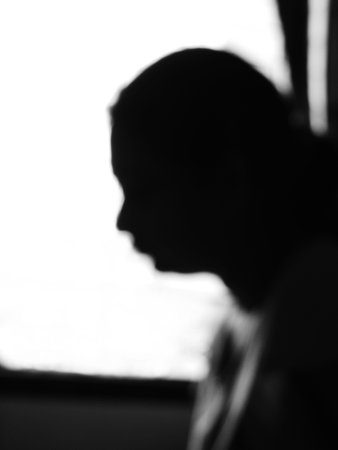Blurred out black & white sideways head & shoulder silhouette portrait of an isolated girl with combed hair sitting at a window and looking out engaged in deep thinking with white blown out backgroundの写真素材