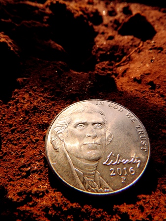 Macro close up of US five cents copper nickel coin in bottom right corner with Thomas Jefferson portrait &amp; liberty 2016 embossed on it, placed on moist red brown soil under twilight sunlight &amp; shadowの写真素材