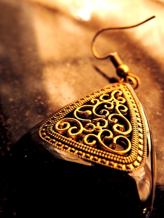 macro close up of a large triangular Mughal style cut out earrings with thick black enamel border and intricate golden curved carving with small golden dots around it placed on a brown surfaceの写真素材