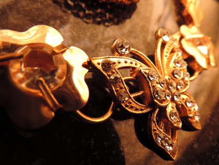 warm close up image of glittering gold jewelry of diamonds studded golden butterfly with gold flowers and petals on either sides kept diagonally placed on dark surface with light reflections visibleの写真素材