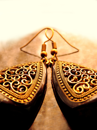 close up of a pair of large triangular Mughal style cut out earrings with thick black enamel border and intricate golden curved carving shot from front placed in symmetry on a golden brown surfaceの写真素材