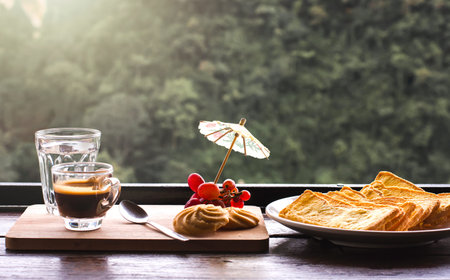 Hot coffee on wooden table with cookies, toast and red grapes. Blurred mountain view background.