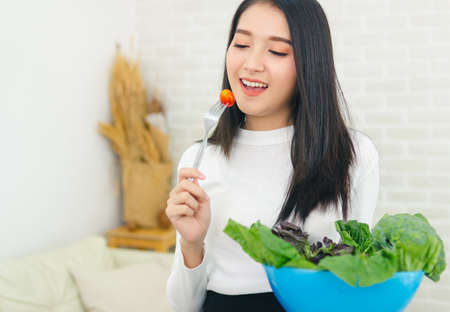asian beauty woman stand holding fork with tomato and holding salad bowl in house.の写真素材