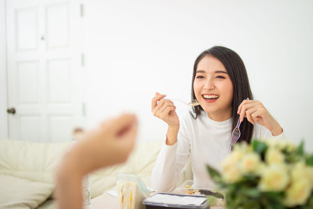 Two beautiful Asian women are sitting and eating new normal lifestyle couple friend use plastic spoon and fork eat foodの写真素材