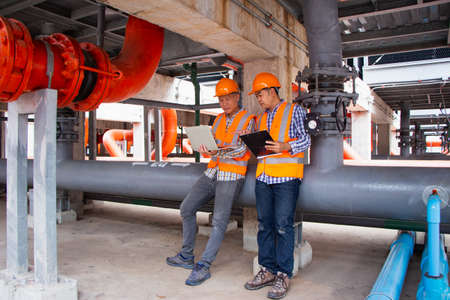 Asian male engineer inspecting pipe fittings and chillers, refrigeration plant refrigeration plant in  factory. men mechanics wear helmets and Reflective Safety Vests, use laptops, notebooks, and walkie-talkies work in factories with pipes background.の写真素材