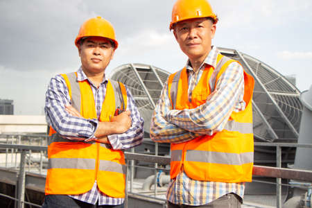 Asian male engineer inspecting pipe fittings and chillers, refrigeration plant refrigeration plant in  factory. male mechanic wearing helmet and Reflective Safety Vest uses laptop, tablet, equipment with sets of cooling towers in conditioning systems background.の写真素材