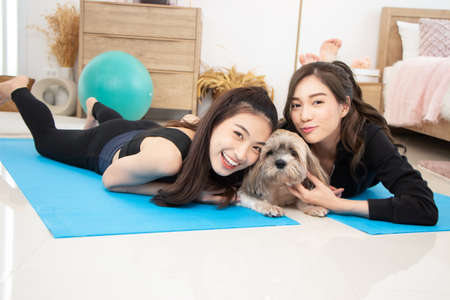 Two beautiful Asian girls wearing workout clothes and smiling with dogs in bedroom. woman on exercise mat with dumbbells and tabletの写真素材