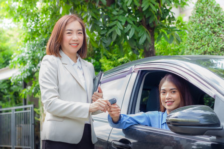 Beautiful young woman car salesman holding car keys and handing it to Asian female customerの写真素材