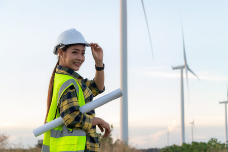 Female engineer wearing safety helmet and holding blueprints with wind turbines backgroundの写真素材