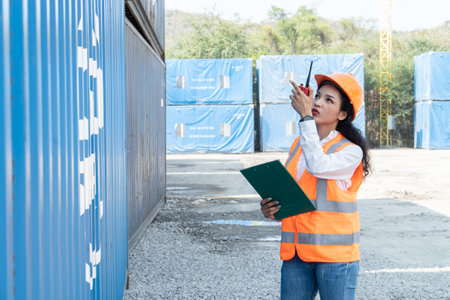 Female engineer holding clipboard and talking on walkie talkie in front of containerの写真素材