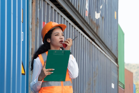 Portrait of Asian woman engineer holding clipboard and talking on mobile phone while standing in container yardの写真素材