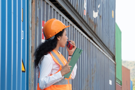 Portrait of female worker holding clipboard and talking on mobile phone while standing at container yardの写真素材