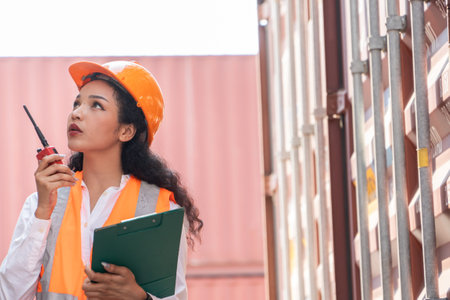 Portrait of female engineer holding clipboard and walkie talkie while standing near container yardの写真素材