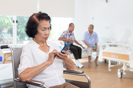 Elderly woman using mobile phone while patient sitting in hospital wardの写真素材