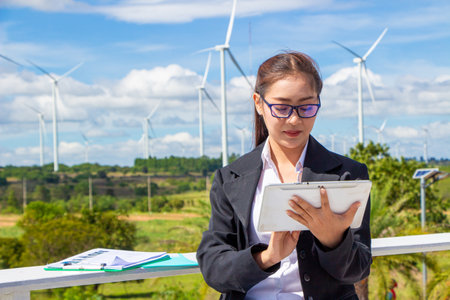 Energy engineer female working at wind farm. asian business woman wearing suit holding tablet and clipboard with windmill environment renewable energy.の写真素材