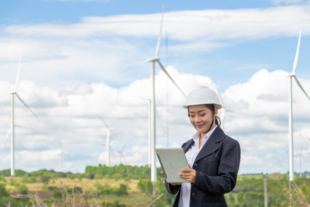 Energy engineer female working at wind farm. asian business woman wearing suit holding tablet and clipboard with windmill environment renewable energy.の写真素材