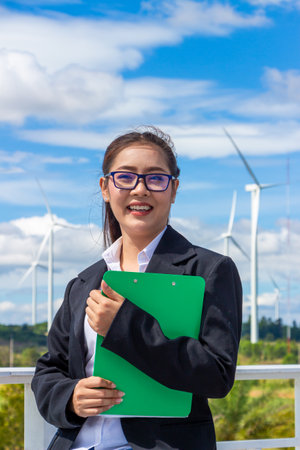 Energy engineer female working at wind farm. asian business woman wearing suit holding tablet and clipboard with windmill environment renewable energy.の写真素材
