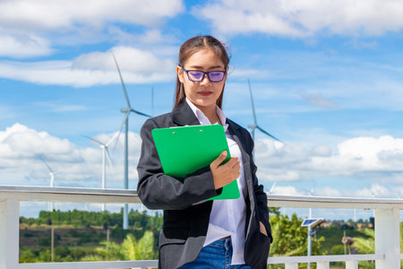 Energy engineer female working at wind farm. asian business woman wearing suit holding tablet and clipboard with windmill environment renewable energy.の写真素材