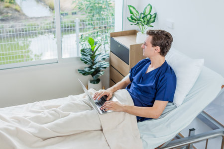 Caucasian patient using laptop in hospital room. man sick patient lying in a hospital bed.の写真素材
