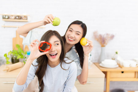Two asian young women eating fresh vegetables in the kitchen at homeの写真素材
