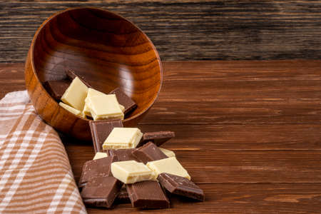 side view of dark and white chocolate pieces scattered from a wooden bowl on rustic background with copy spaceの写真素材