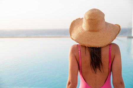 Back view of young woman in pink swimsuit and straw hat standing near swimming poolの写真素材