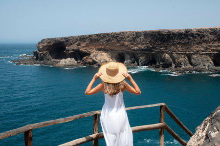 A woman in a white dress and a straw hat stands on the edge of the cliff and looks at the sea