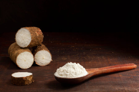 Coconut flour on a wooden spoon and raw cassava on a dark background.