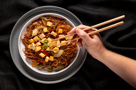 Hands holding chopsticks and soy sauce noodles with tofu and vegetablesの写真素材