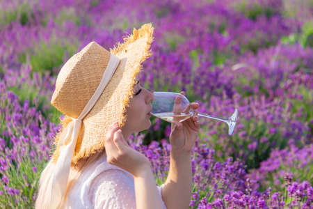 a girl holds a glass of white wine against the backdrop of a lavender field. A girl drinks wine in a lavender field. selective focusの写真素材