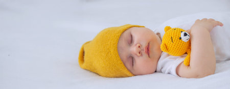 A child in a white bodysuit is sleeping on a white background, hugging a small knitted yellow teddy...
