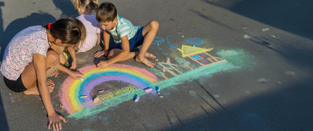 Children draw a picture with chalk on the asphalt. A rainbow, house, sun, clouds are drawn on the...