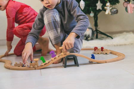 A boy and a girl are playing with a wooden toy train near a Christmas tree at home