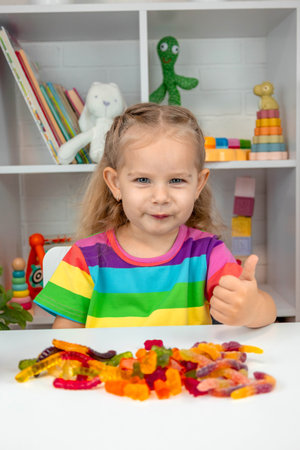 Happy little girl in a rainbow t-shirt playing with and eating colorful gummy candies or vitaminsの写真素材